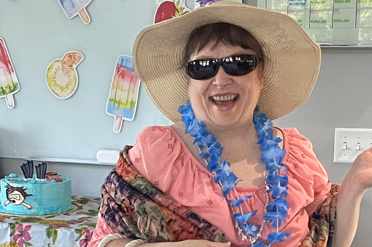 Woman smiling indoors wearing a straw sun hat, black sunglasses, a blue lei, pink top, and a colorful shawl.