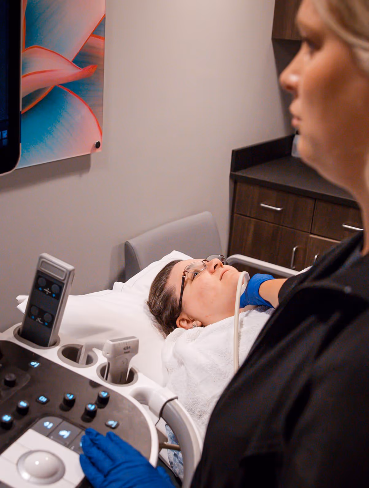 Woman lying on a medical bed receiving an ultrasound scan on her neck from a healthcare professional wearing blue gloves.