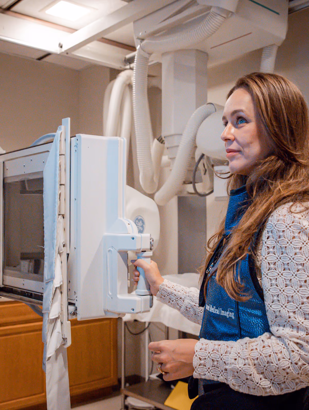 A medical professional wearing a blue protective vest operates an X-ray machine in a clinical room.
