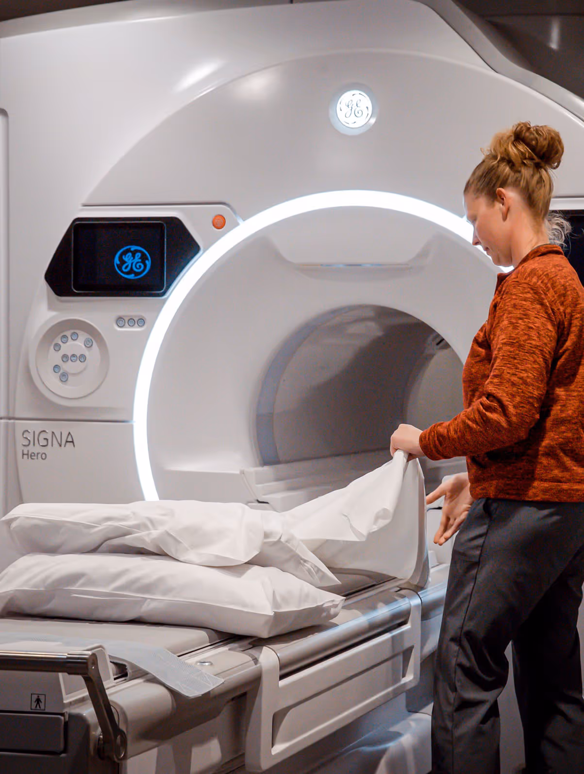 Technician preparing a pillow with white sheets on an MRI machine table inside a medical examination room.