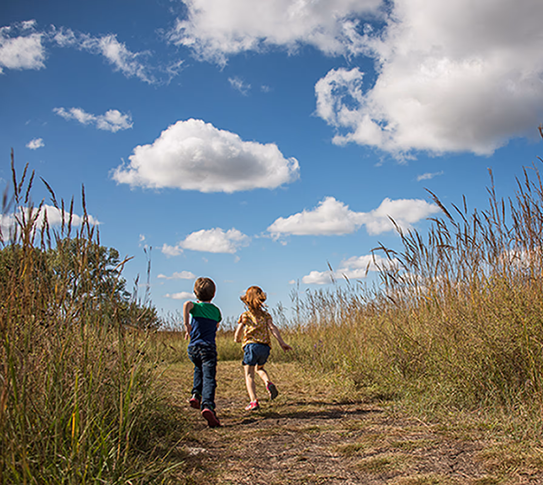 Two children running on a dirt path surrounded by tall grass under a blue sky with scattered clouds.