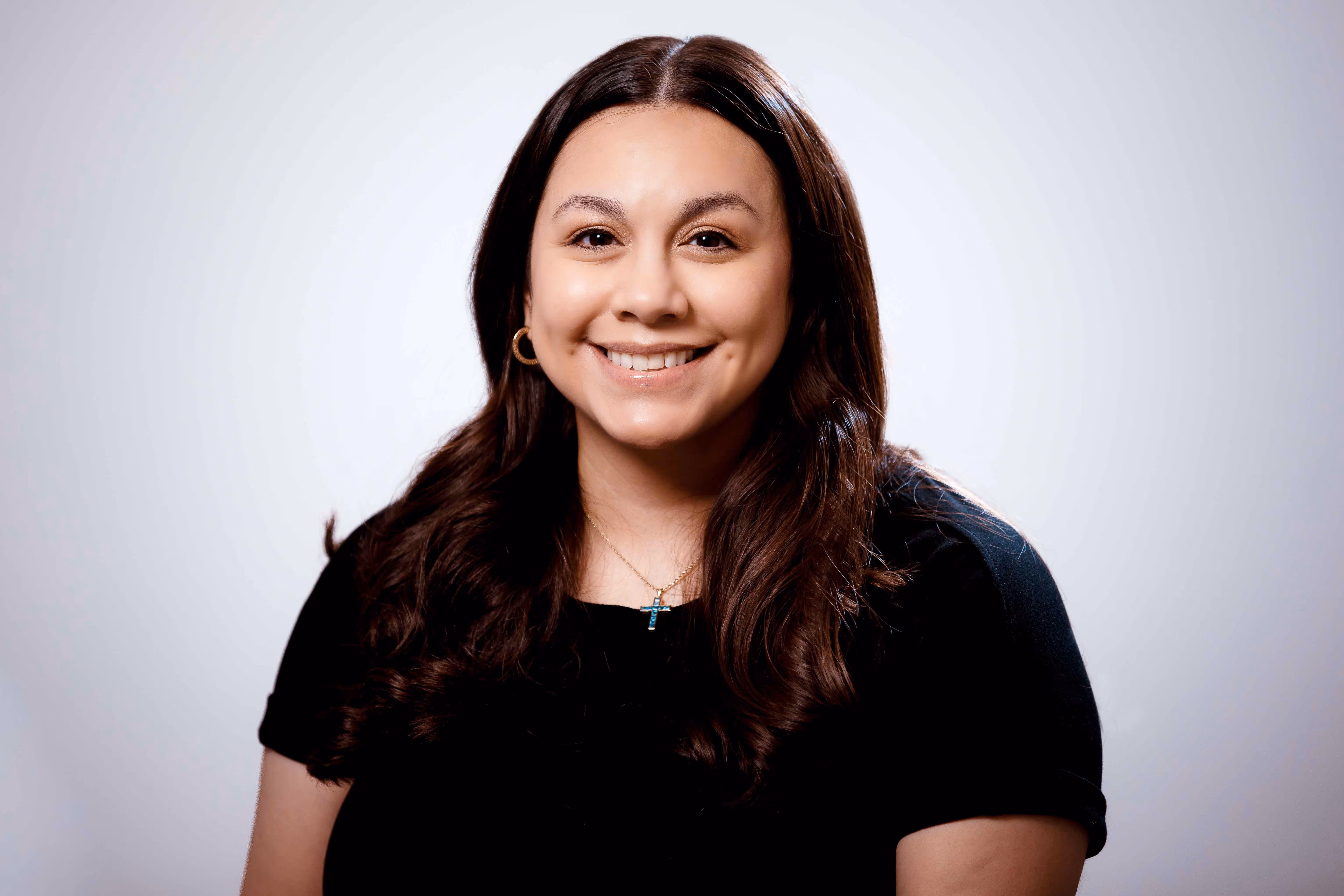 Smiling woman with long dark hair wearing gold hoop earrings, a cross necklace, and a black top against a light gray background.