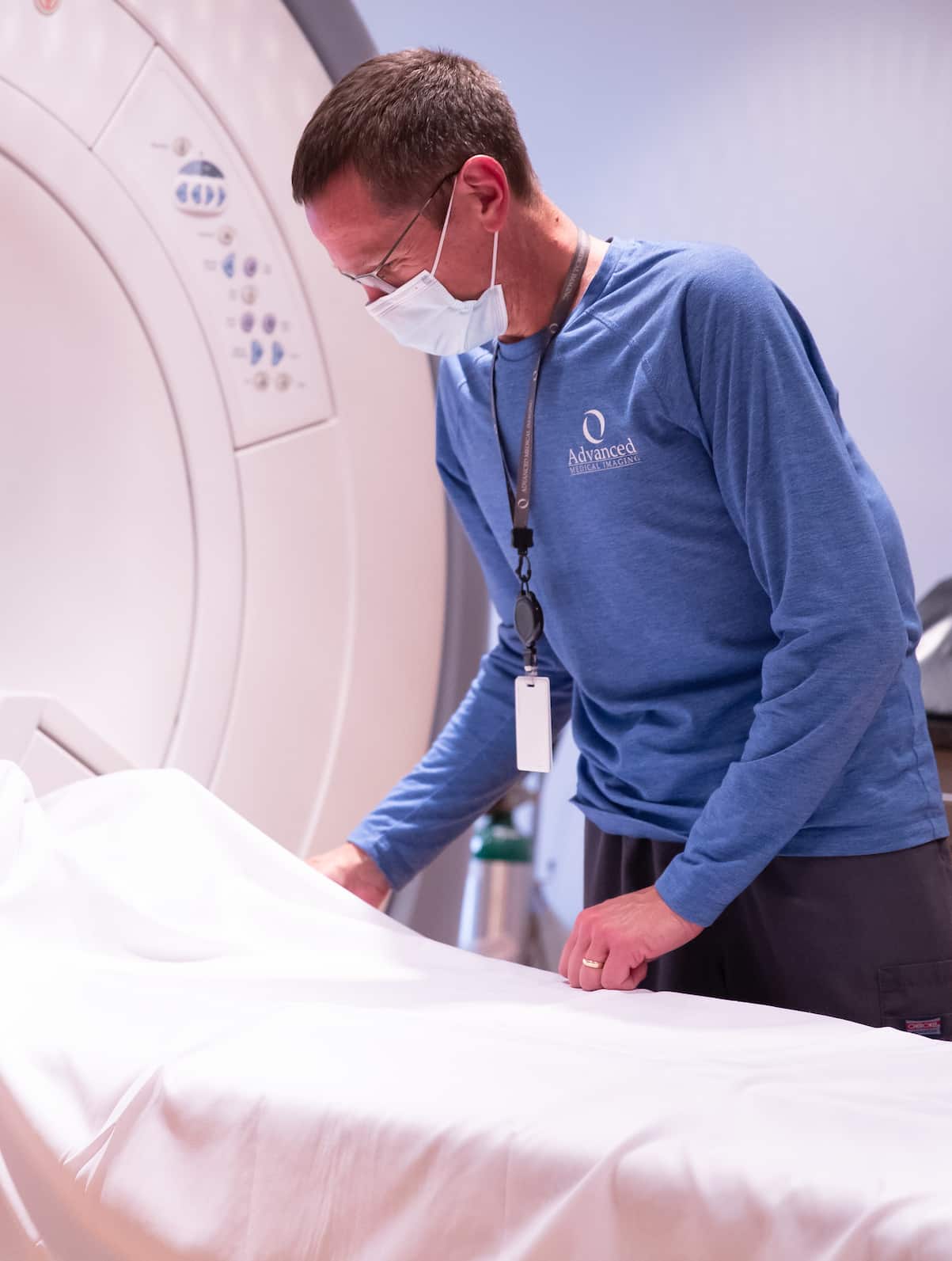 A masked technologist in blue preparing the patient table at the opening of a scanner.