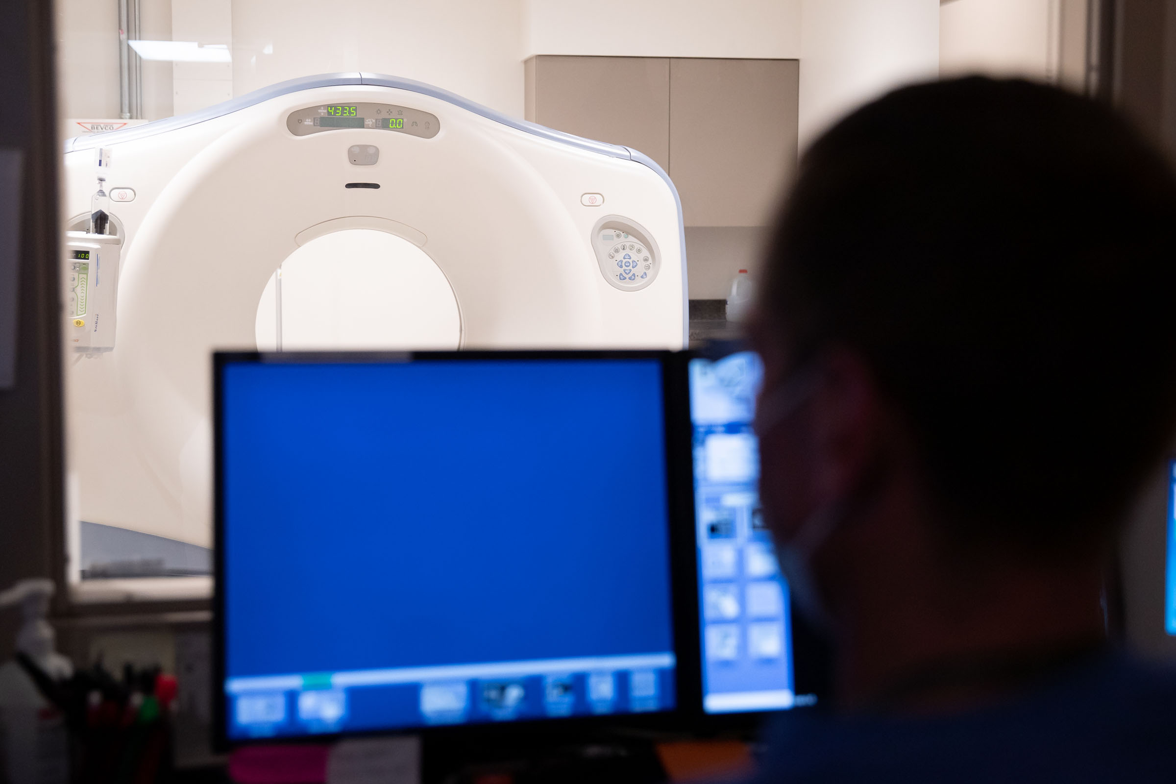 A technologist seated at a monitor in the foreground with a CT scanner in the room behind.