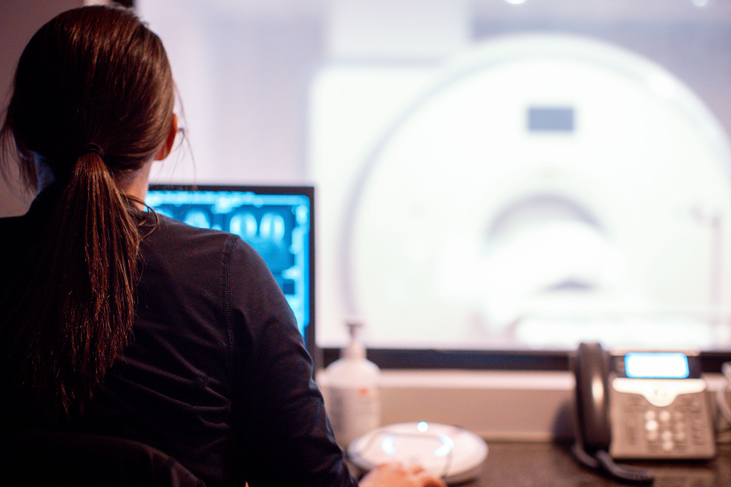 A technologist at a workstation reviewing scan images, with an MRI scanner visible through the window.