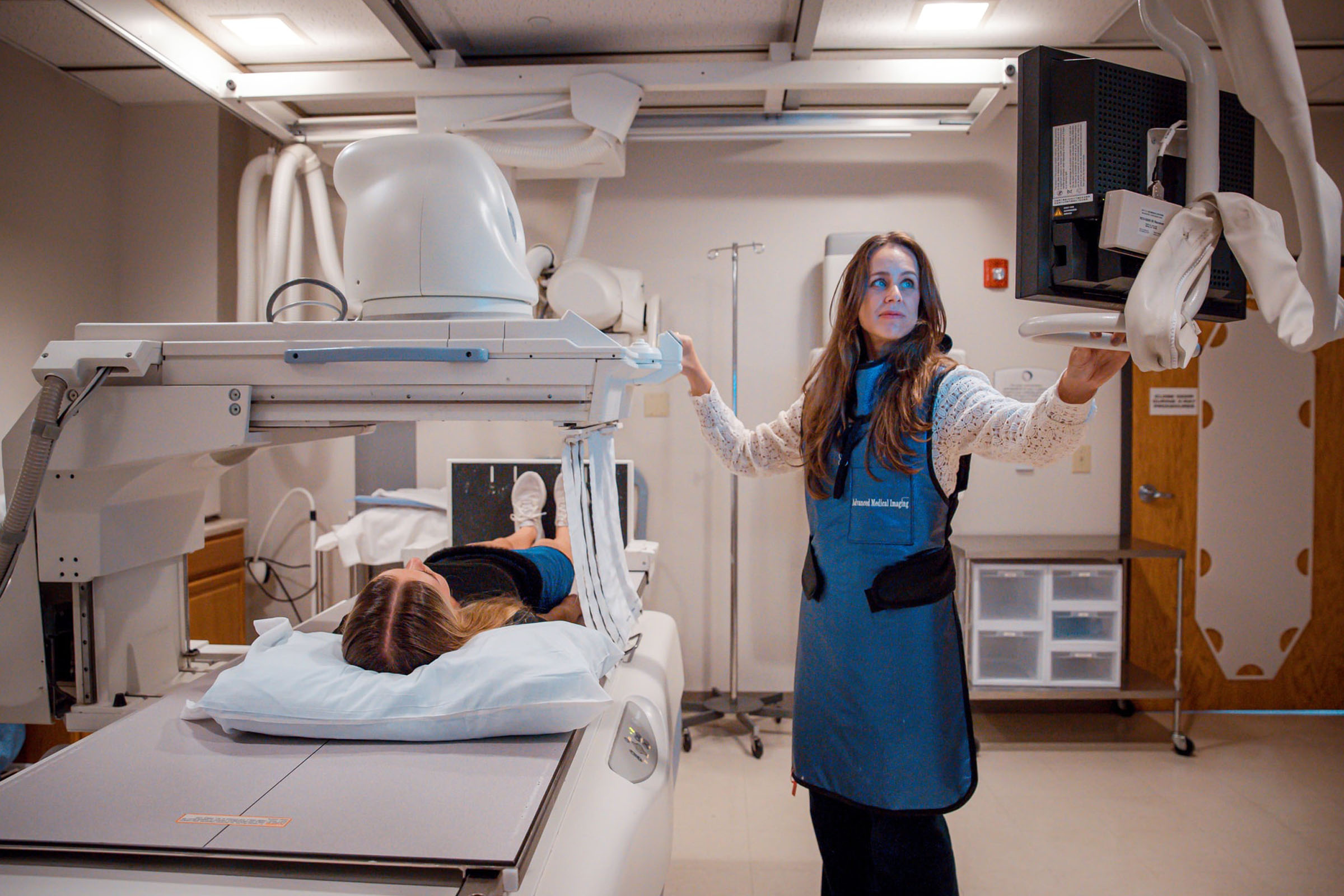 A technologist in a lead apron operating a fluoroscopy unit over a patient on the exam table.