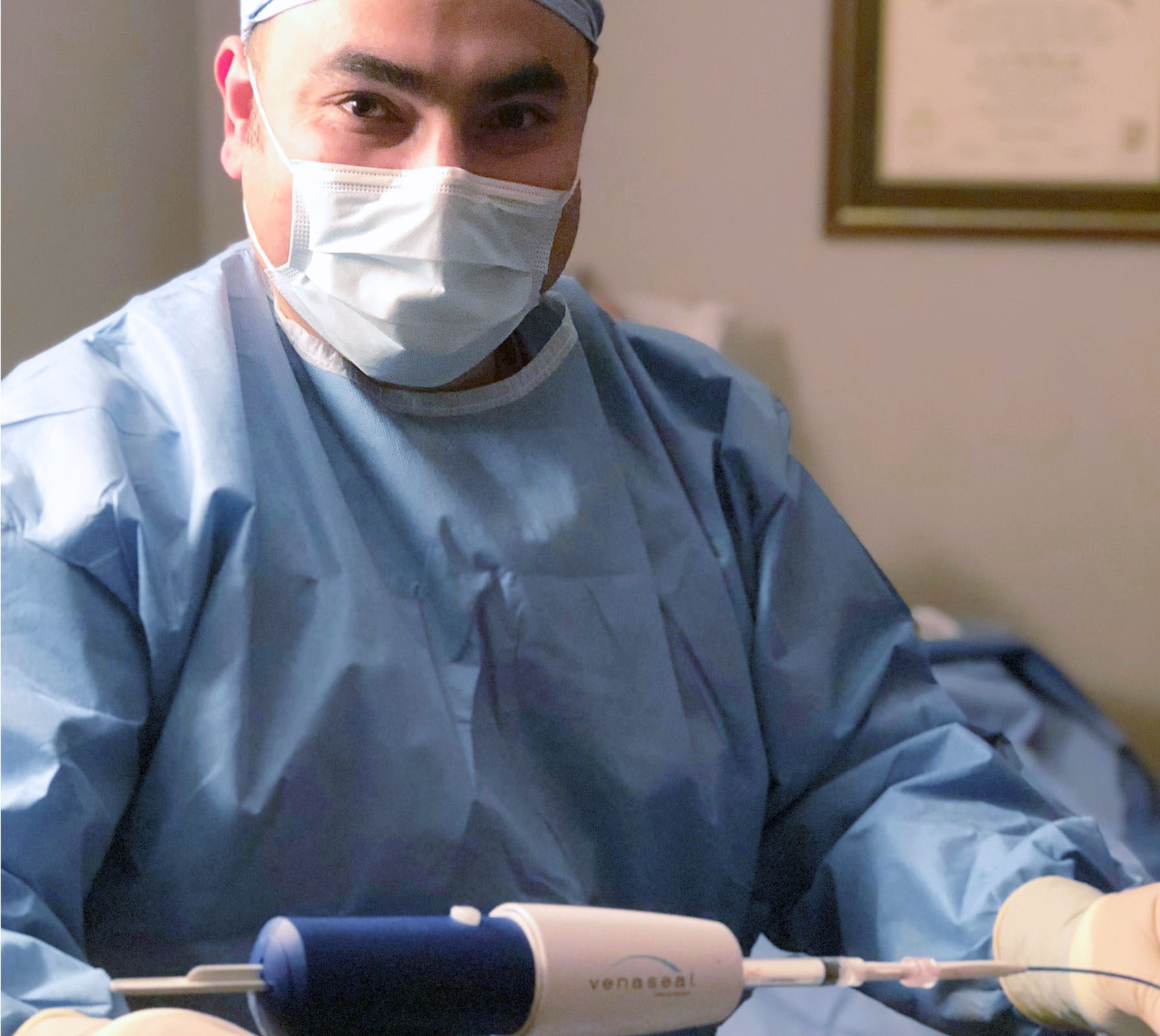 A masked physician in surgical scrubs operating a vein treatment device.