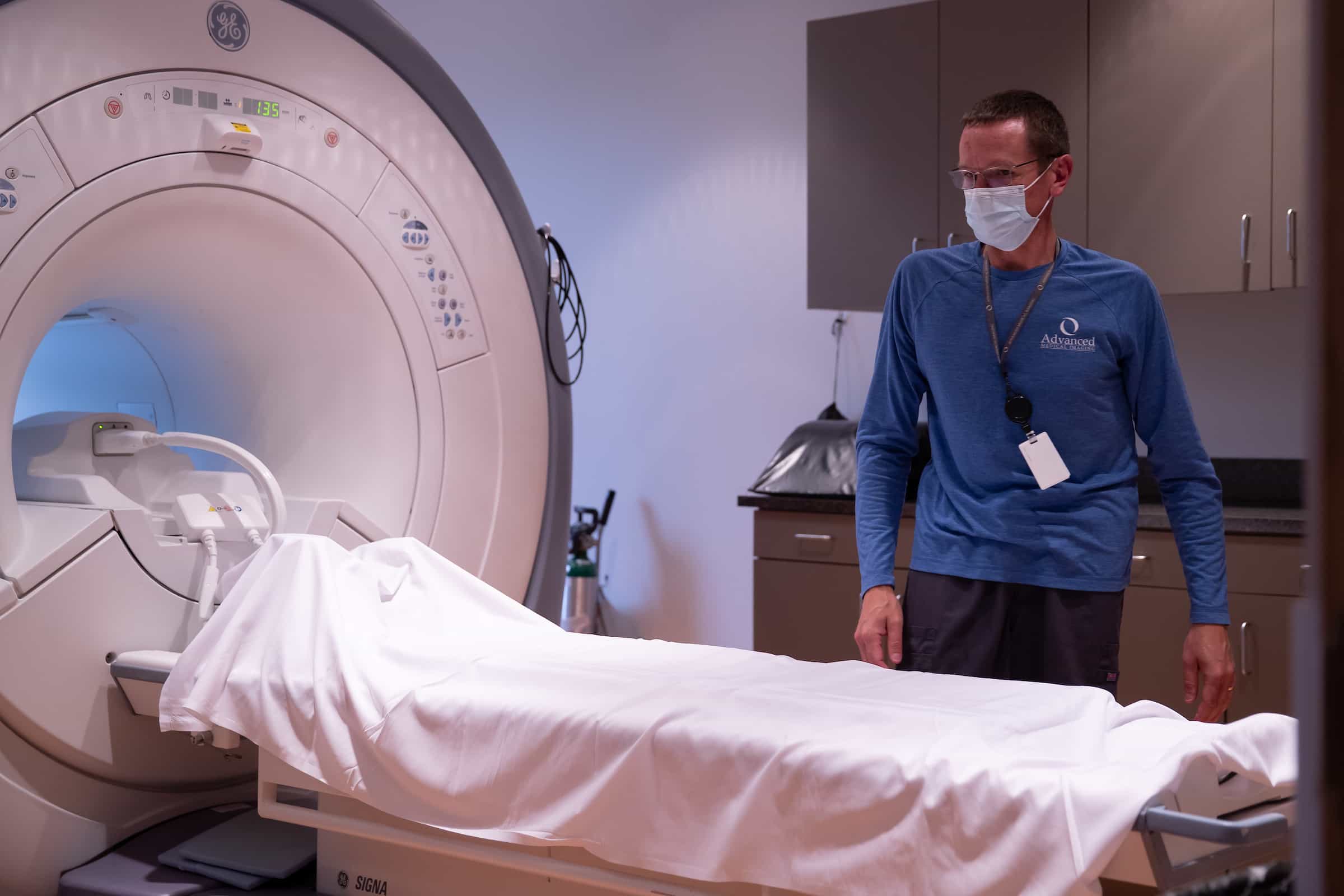 A masked technologist in blue standing beside an MRI scanner with the patient table prepared.