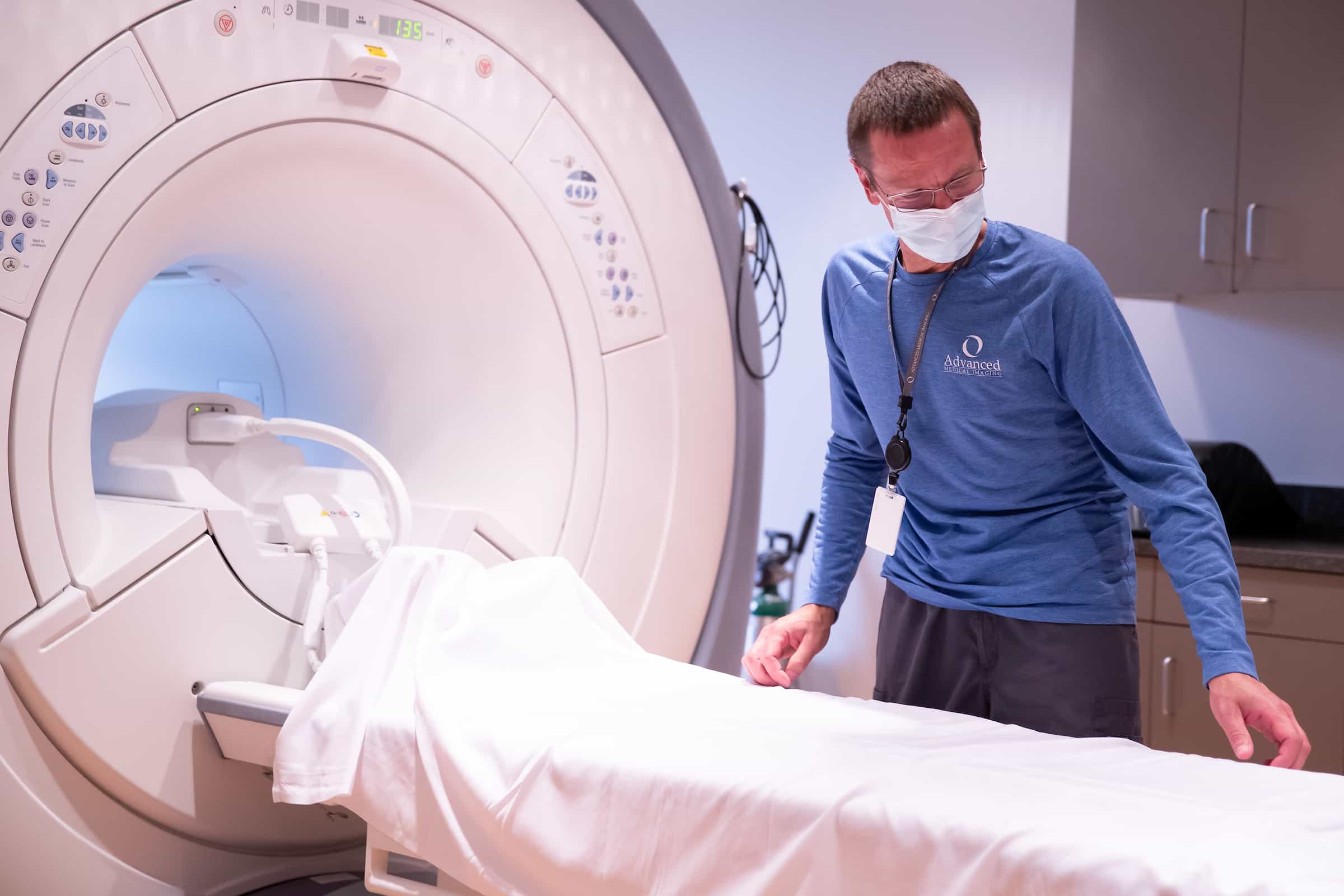 A masked technologist in blue preparing the patient table at the opening of an MRI scanner.