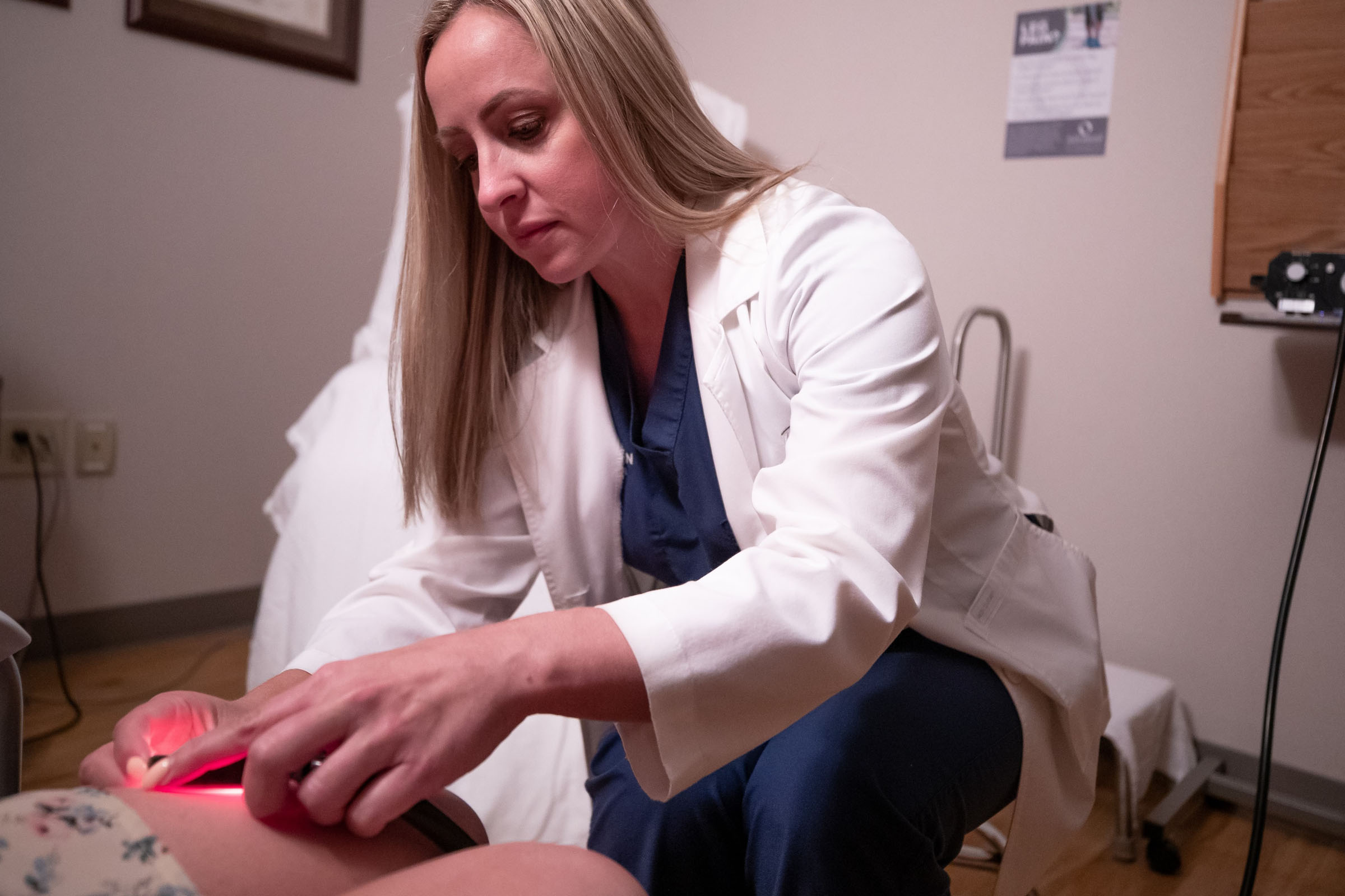 A clinician in a white coat treating a patient's leg with a handheld red light device.