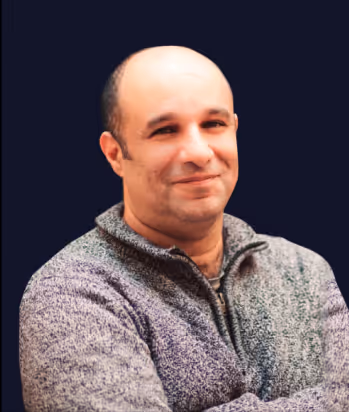 Smiling bald man wearing a gray sweater with arms crossed against a dark background.
