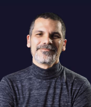 Middle-aged man with short dark hair and salt-and-pepper beard wearing a dark gray turtleneck sweater, smiling gently against a dark background.