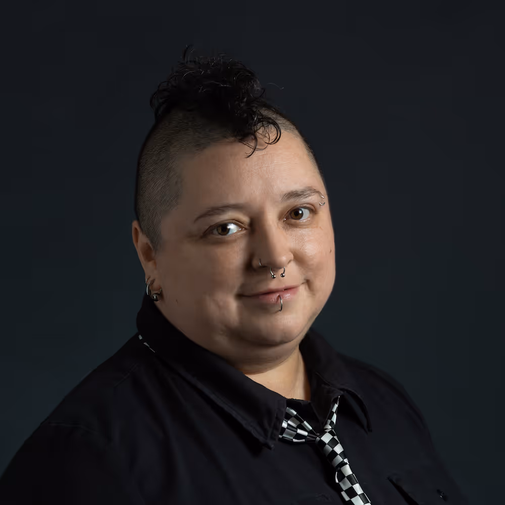 Person with a curly mohawk, facial piercings, and a checkered tie over a black shirt, smiling against a dark background.