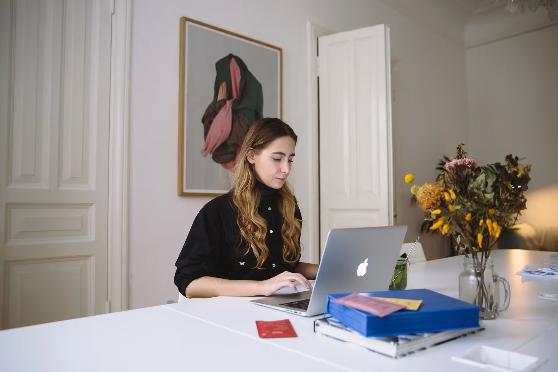Woman with long hair sitting at white desk working on a MacBook laptop in a room with white walls, a framed abstract artwork, and a vase of flowers.