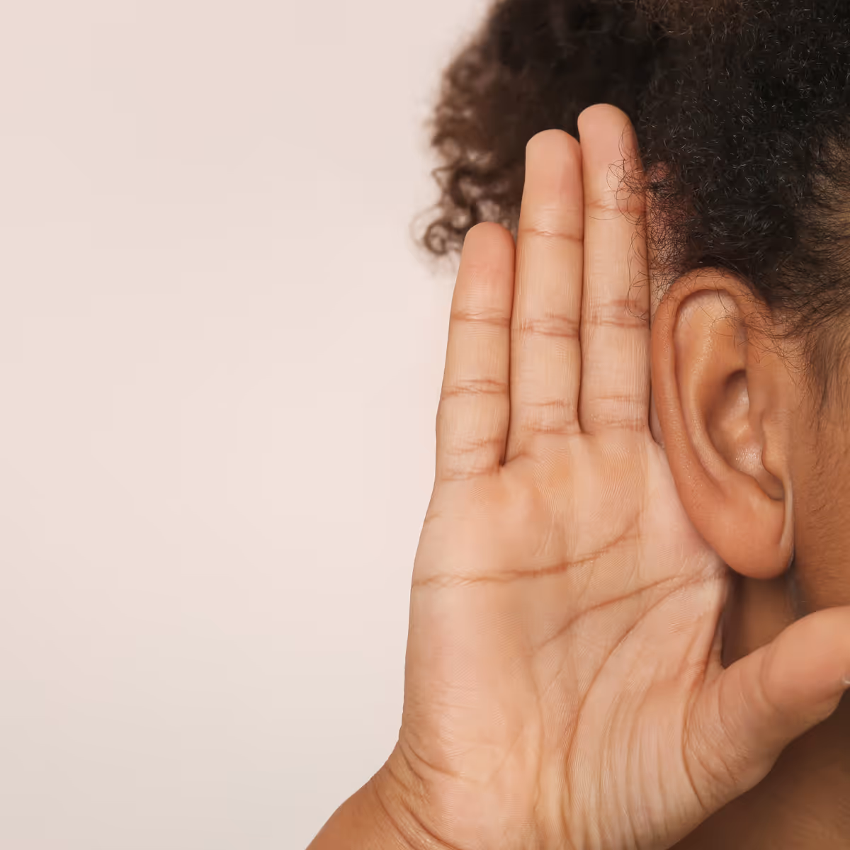 Close-up of a person with curly hair holding their hand behind their ear to listen closely.