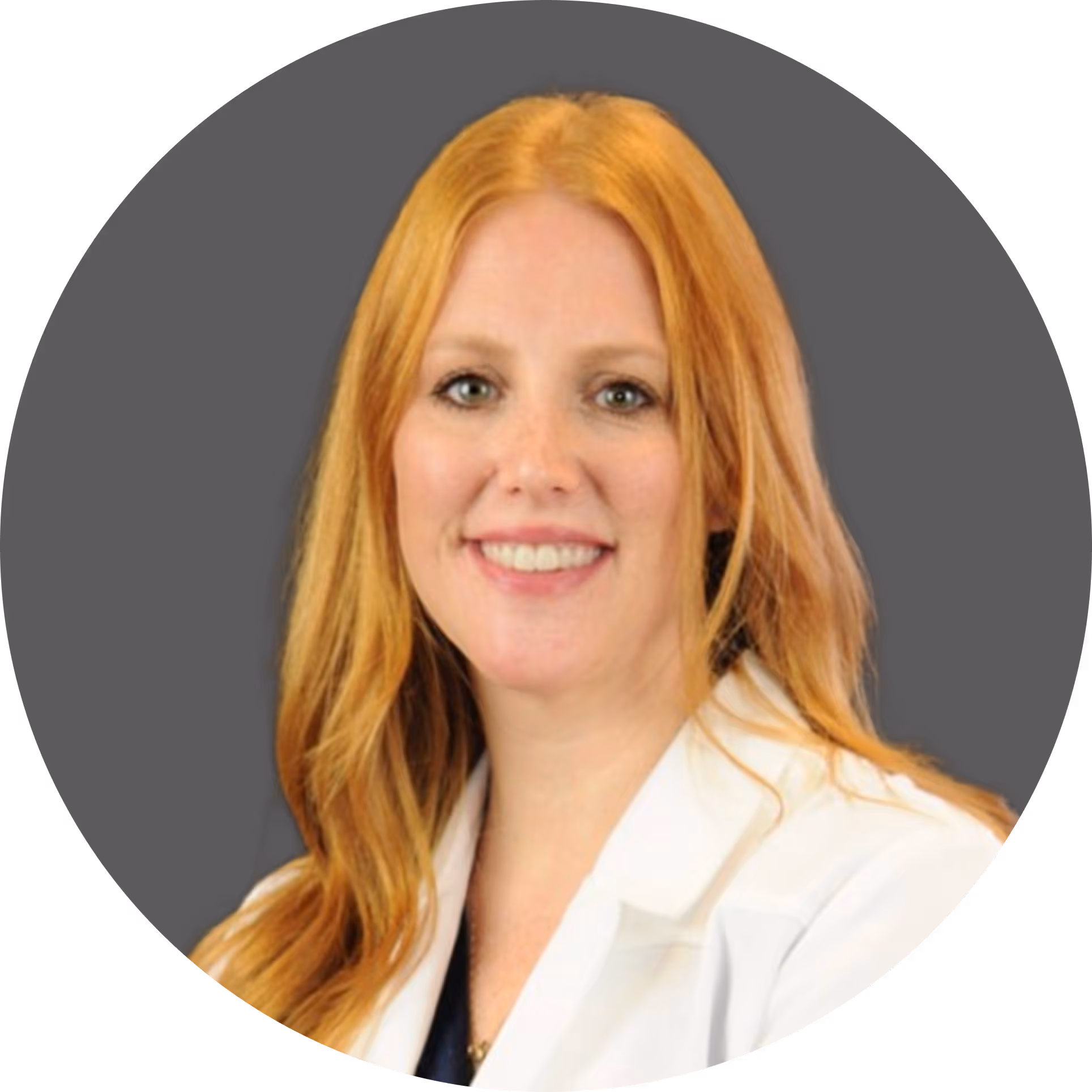 Portrait of a smiling woman with long red hair wearing a white lab coat against a gray background.