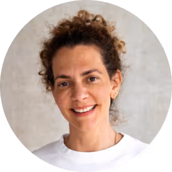 Smiling woman with curly brown hair pulled back, wearing a white top against a light gray background.