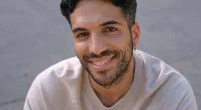 Smiling man with short dark hair and beard wearing a light beige shirt against a neutral background.