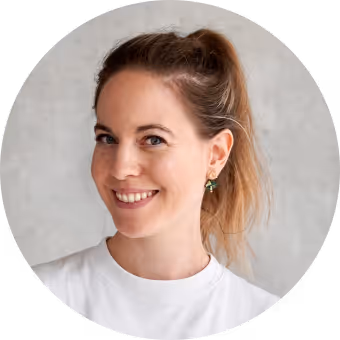 Smiling woman with light brown hair tied back wearing a white shirt and green earrings against a light gray background.