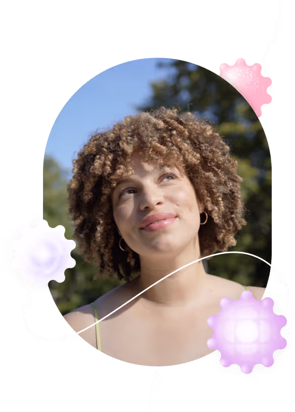 Young woman with curly hair smiling and looking up outdoors with trees and blue sky in background.