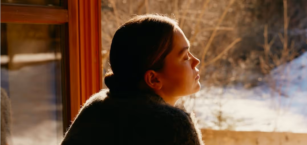 Woman with eyes closed, basking in sunlight by a window overlooking a snowy outdoor scene.