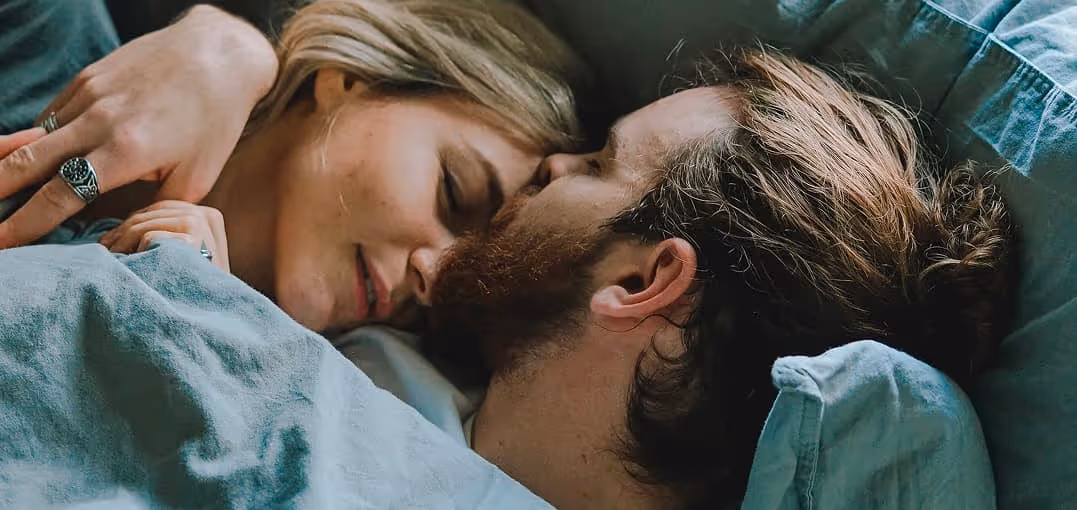 Couple lying in bed close together, eyes closed, sharing an intimate moment under a gray blanket.