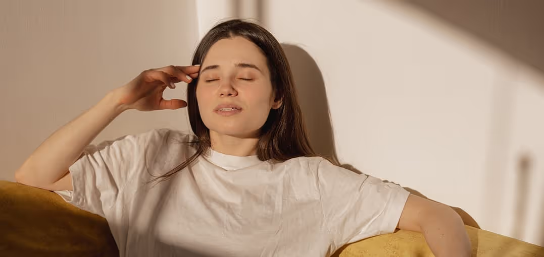 Young woman with closed eyes resting on a yellow couch, touching her temple as if experiencing discomfort.