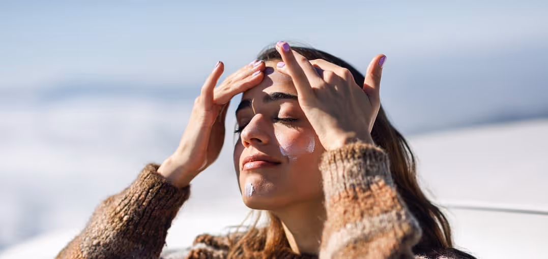 Woman with eyes closed applying sunscreen on her face outdoors in sunlight.