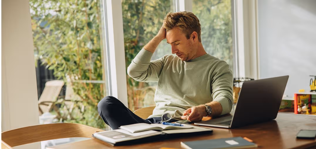 Man sitting at a wooden table in front of a laptop, looking stressed with his hand on his head.