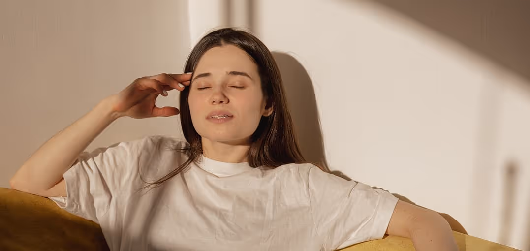 Young woman with closed eyes resting on a yellow couch, touching her temple in soft sunlight.