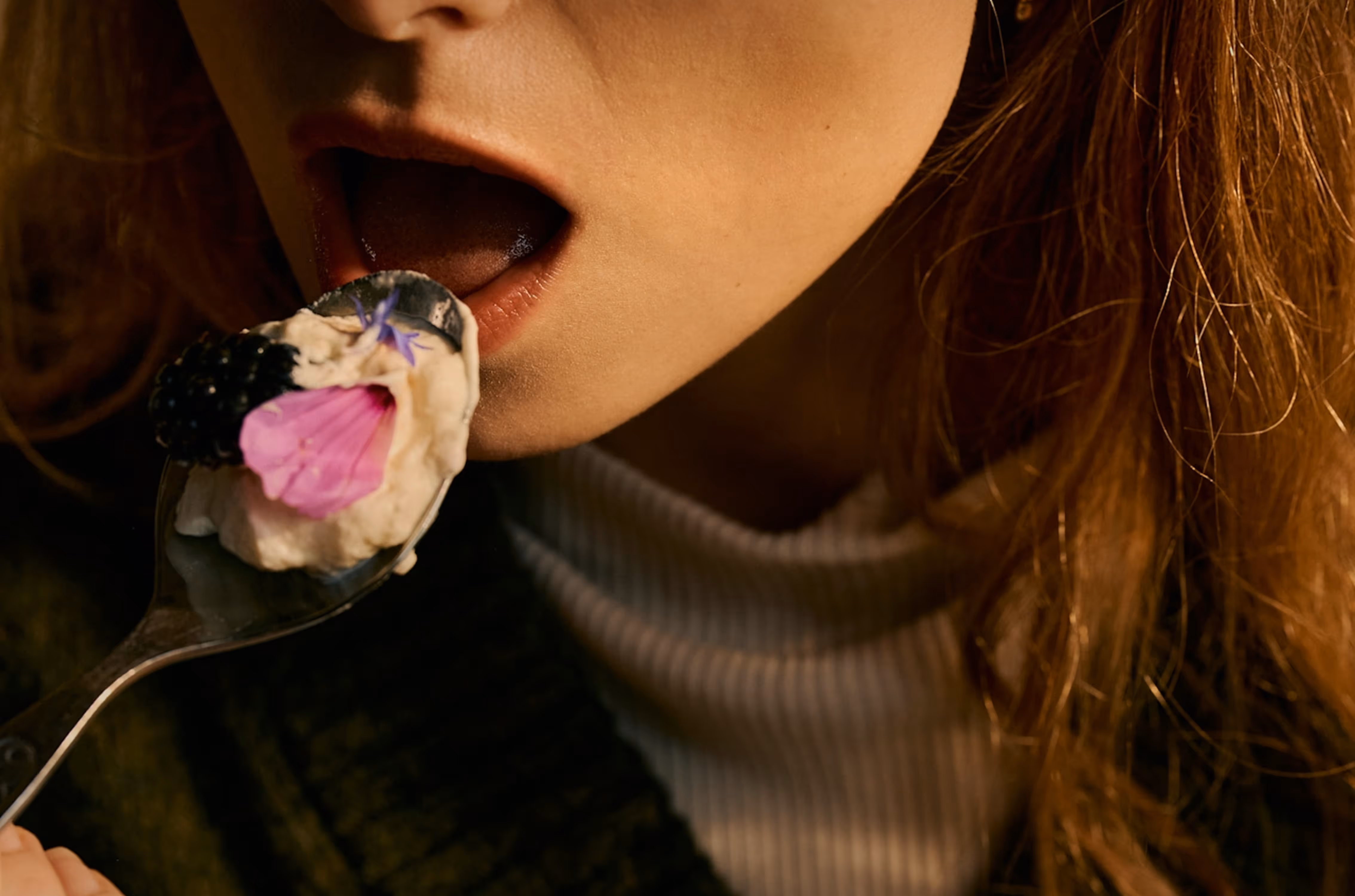 Close-up of a woman about to eat a spoonful of creamy dessert topped with a blackberry and edible flowers.