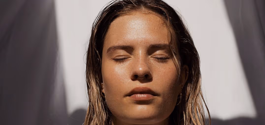 Close-up of a woman with wet hair and closed eyes standing in sunlight with shadows in the background.