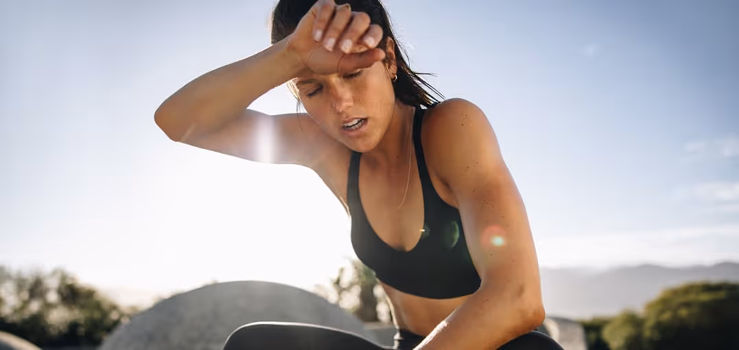 Tired woman in workout clothes wiping sweat from her forehead outdoors in bright sunlight.