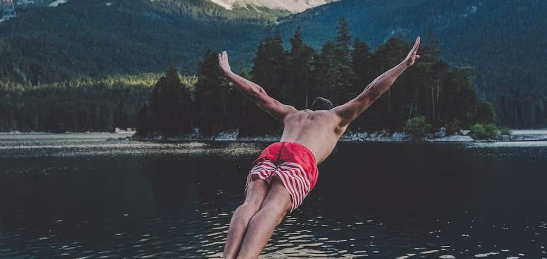 Man diving into a lake surrounded by forested mountains, wearing red and white swim trunks.
