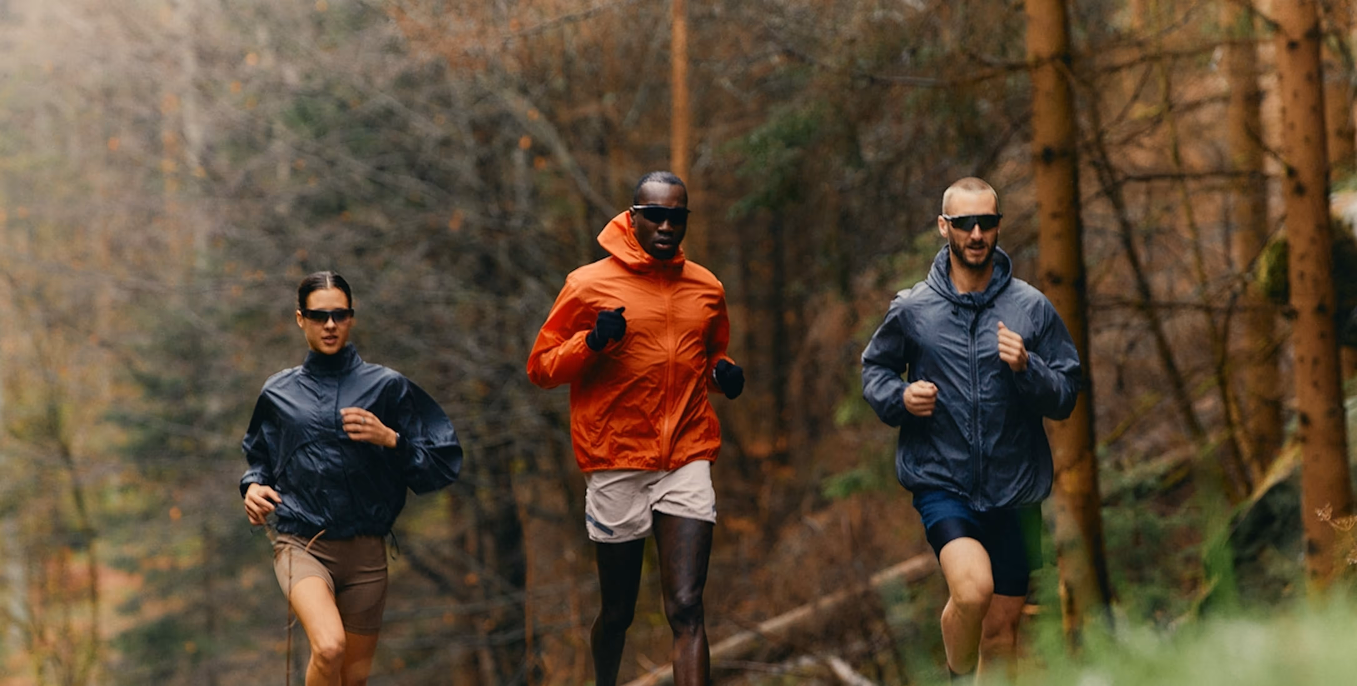 Three runners wearing jackets and sunglasses jogging on a forest trail.
