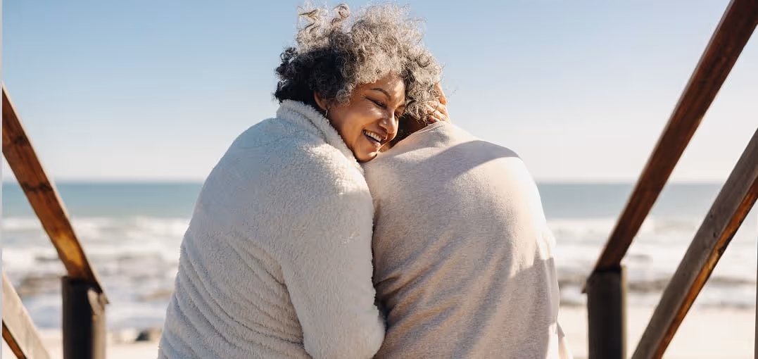 Elderly couple hugging and smiling on a wooden deck overlooking the ocean.