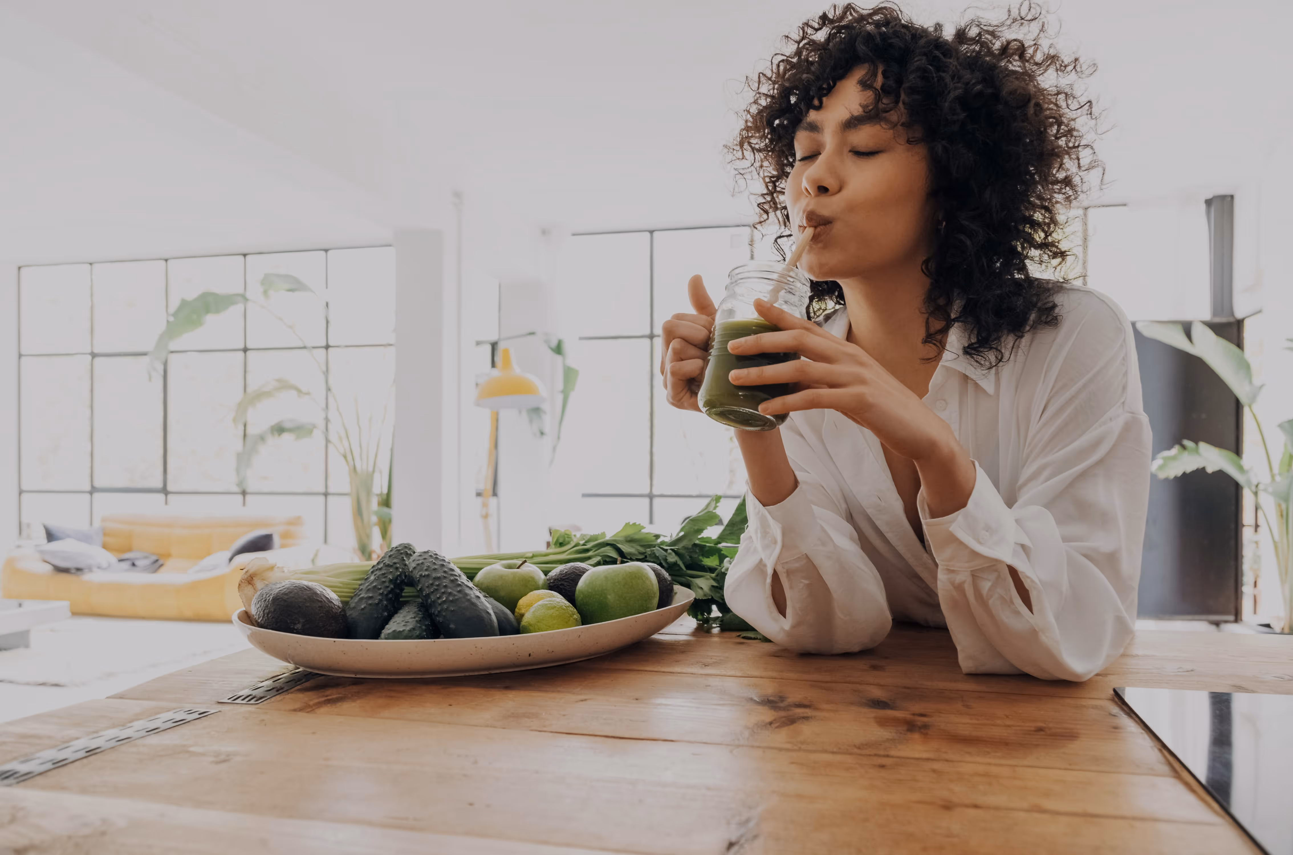 Woman with curly hair in white shirt drinking green smoothie from a glass jar at a wooden table with a plate of fresh vegetables and fruits.