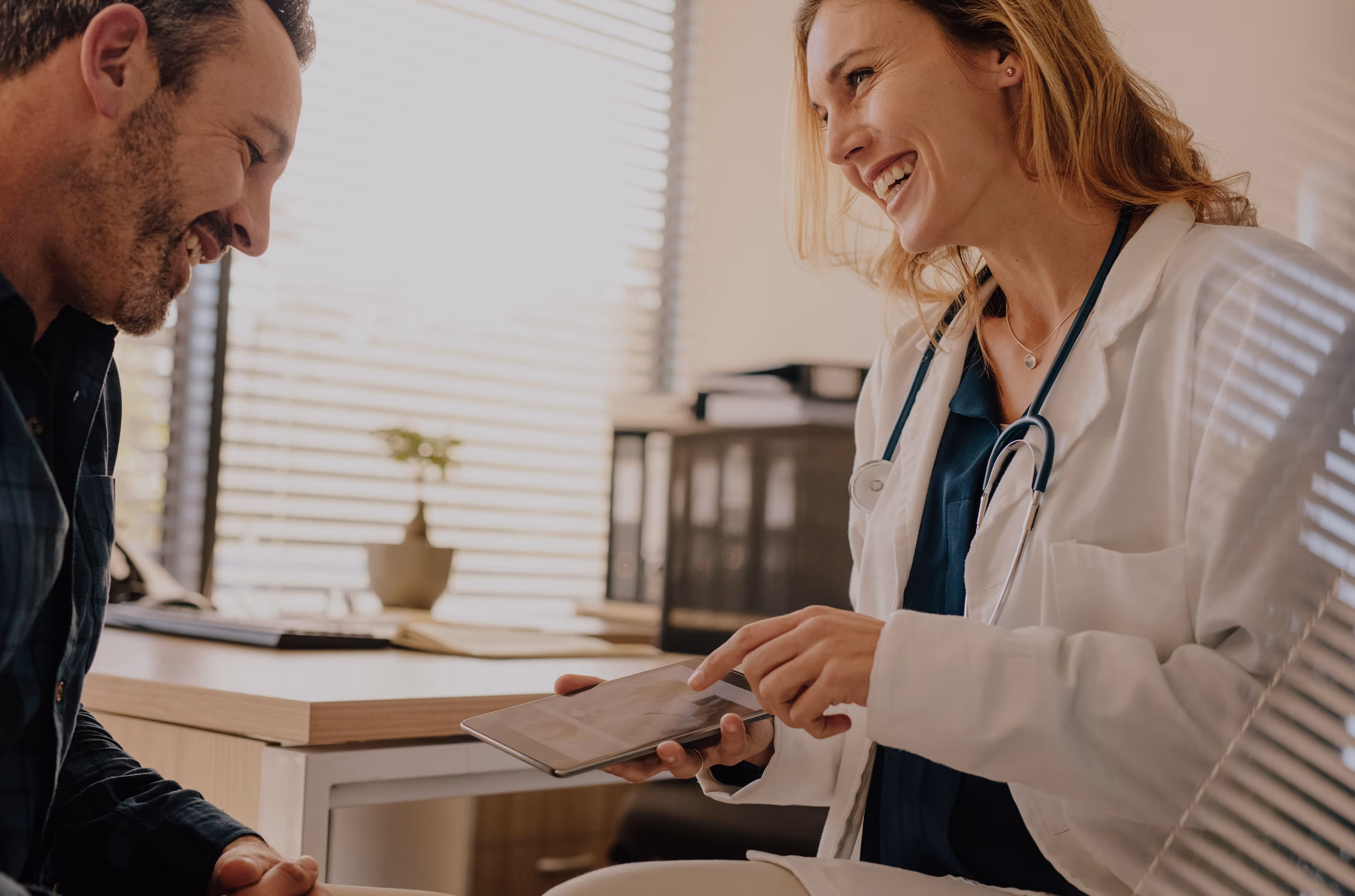 Female doctor smiling and showing information on a tablet to a smiling male patient in a bright office.