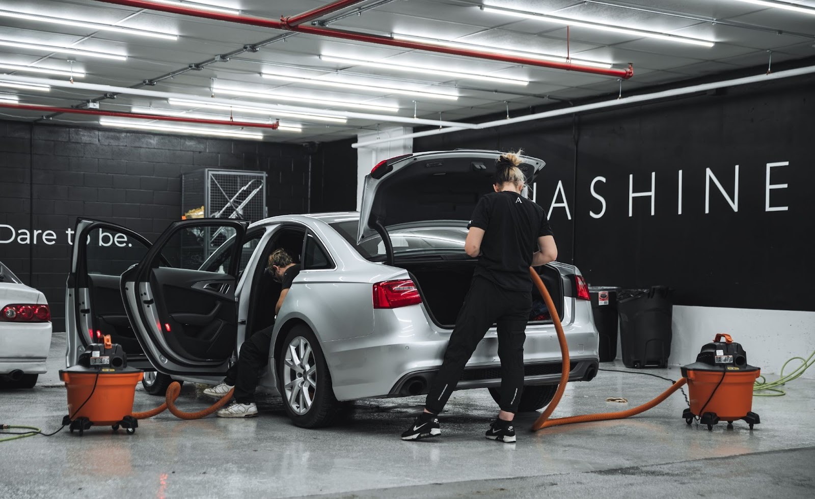 Two car detailers vacuuming and cleaning the interior of a silver sedan inside a professional detailing bay with bright lighting and “ALPHASHINE” branding on the wall.