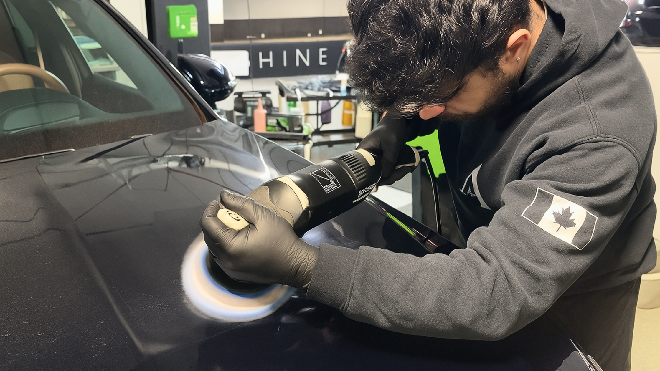 An ALPHASHINE detailing professional wearing black gloves carefully polishes the hood of a dark blue luxury SUV with a dual-action buffer, restoring a deep, reflective shine inside a clean detailing bay.