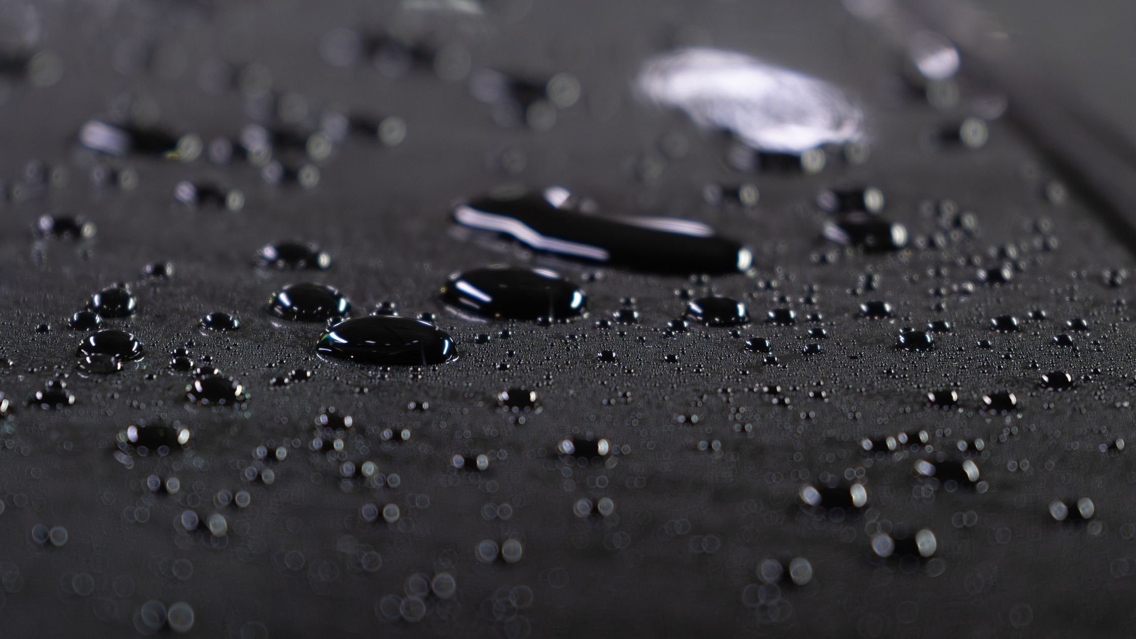 Close up of water droplets beading on a dark car surface, highlighting a smooth, glossy finish with shallow depth of field.