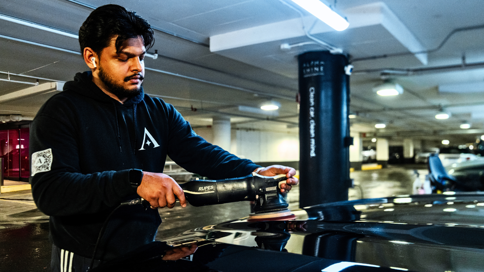 An ALPHASHINE car detailer wearing a black hoodie uses a powered polisher on the roof of a black vehicle inside a parking garage, with overhead fluorescent lighting reflecting off the glossy surface.