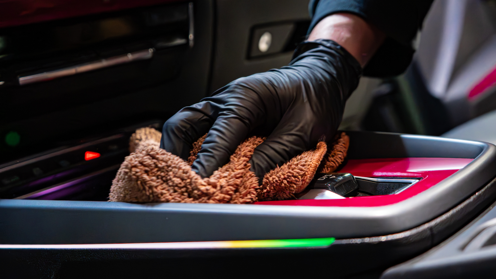 ALPHASHINE car detailer wearing black gloves wipes a car center console with a microfiber cloth, detailing a red and black interior surface.