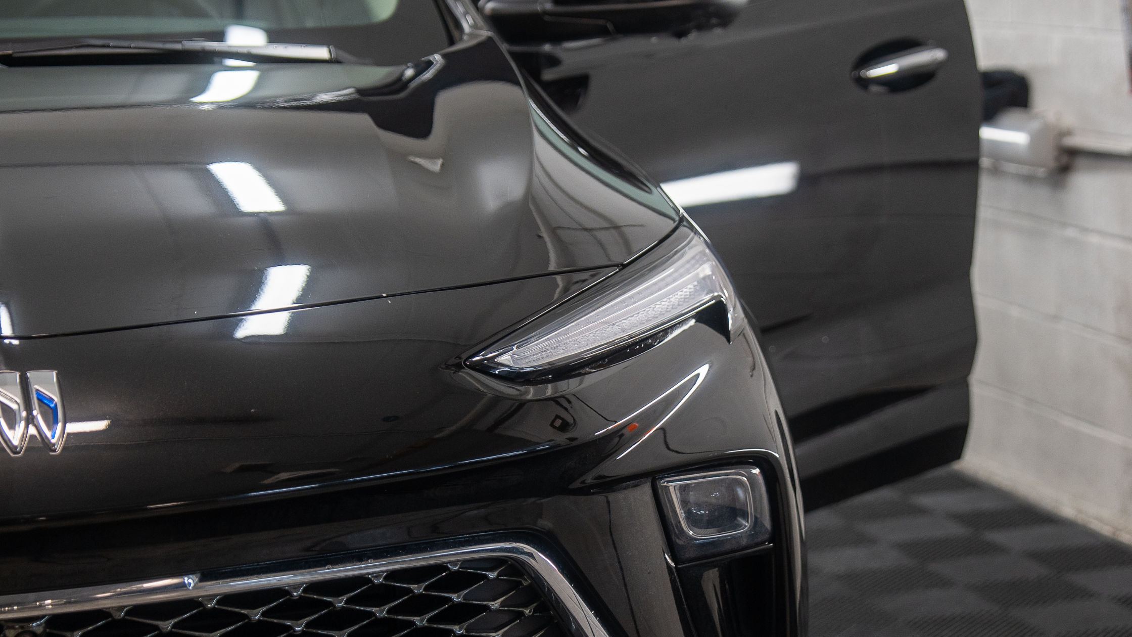 Close up of the front end of a black vehicle inside a garage, showing the headlight, grille, and glossy paint reflecting overhead lights with the driver door open.