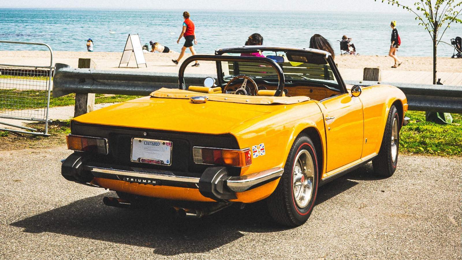 Yellow vintage convertible parked near a lakeside boardwalk, with people walking along the beach in the background on a sunny day.