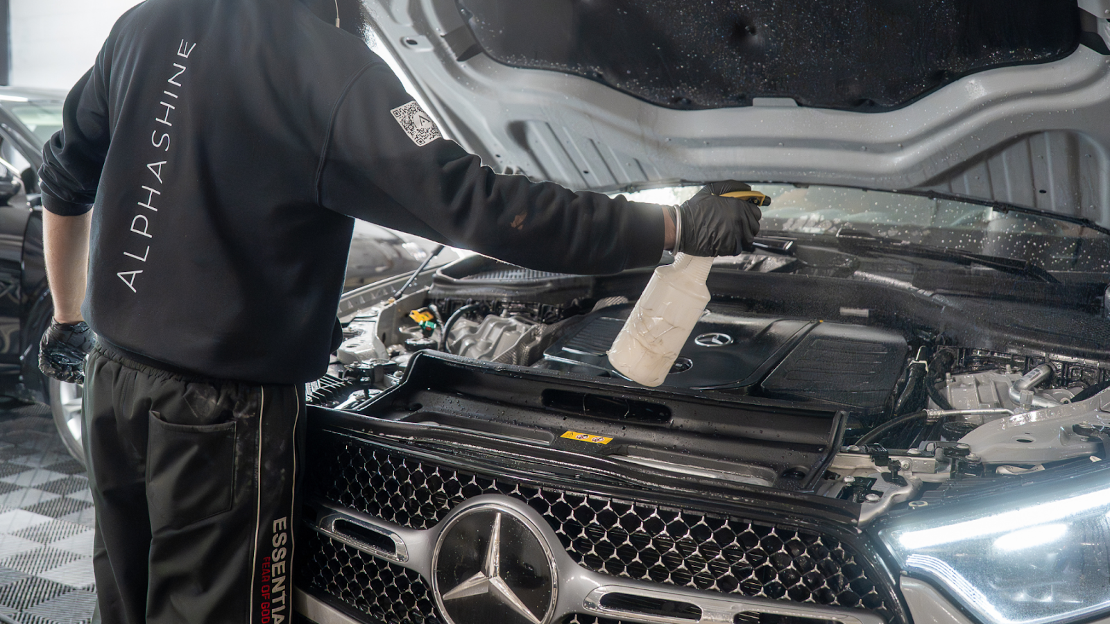 ALPHASHINE detailer wearing black gloves sprays cleaner inside the open engine bay of a Mercedes vehicle in a well lit garage.