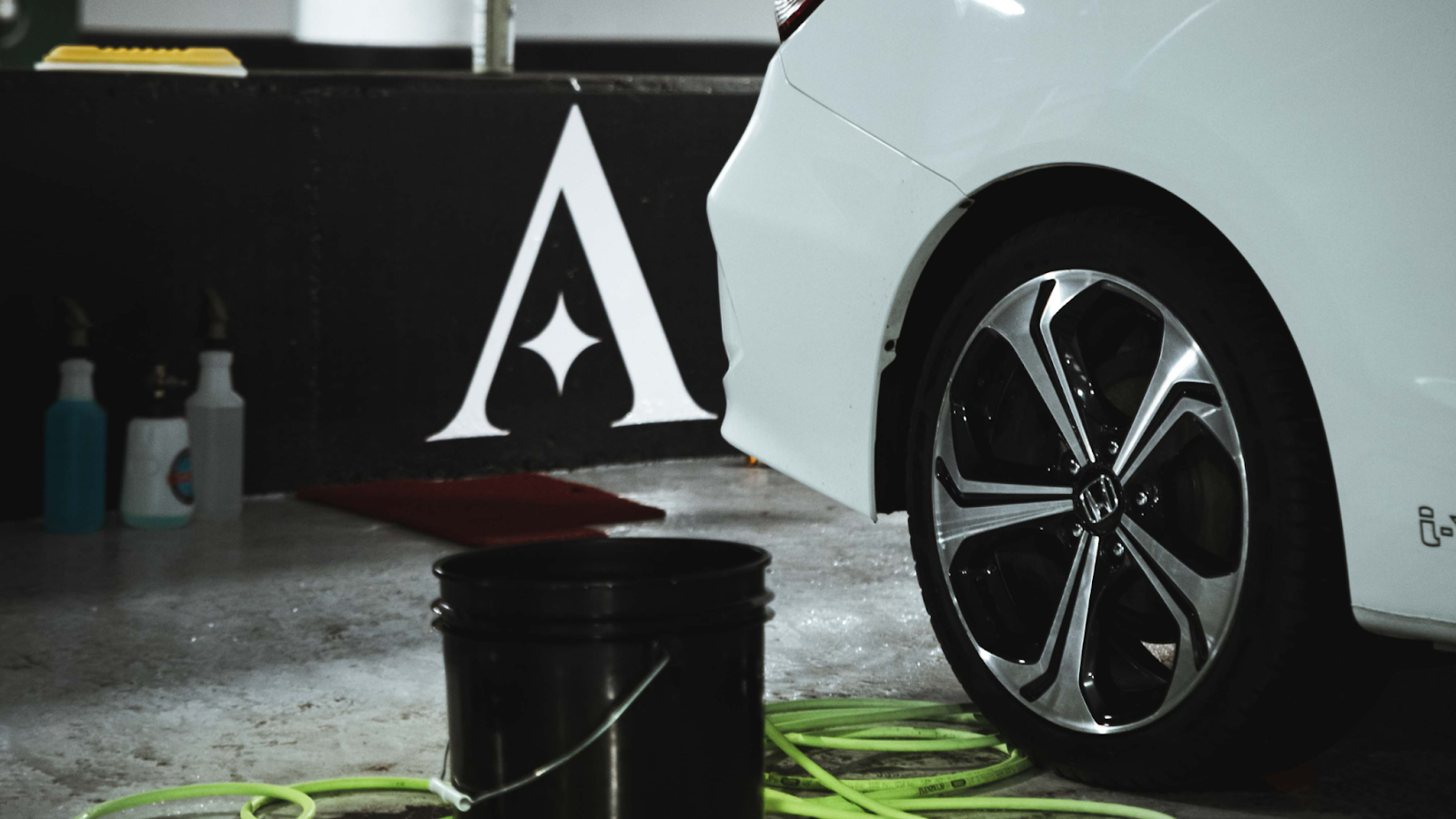 Professional car detailing setup with bucket, hose, and supplies beside a clean white vehicle in a premium auto detailing studio.