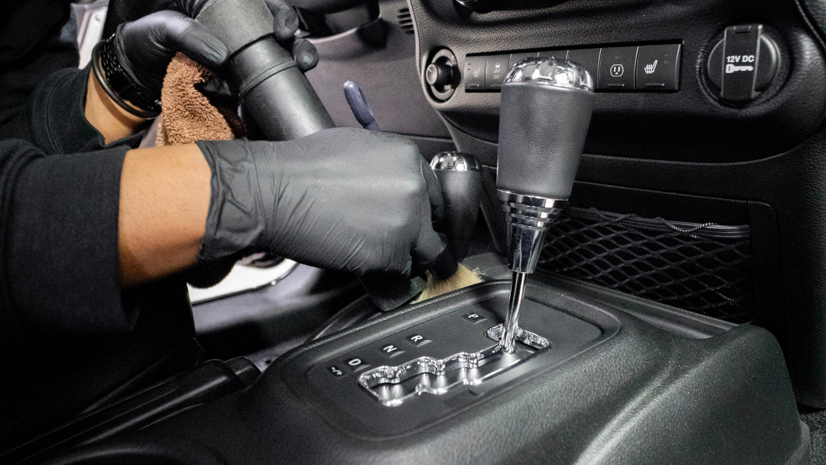 A car detailing technician wearing black nitrile gloves uses a small brush and microfiber cloth to clean around the gear shift and center console of a vehicle. The automatic transmission shifter with chrome accents is in the foreground along with dashboard controls including heated seat buttons and a 12V DC port visible in the background.