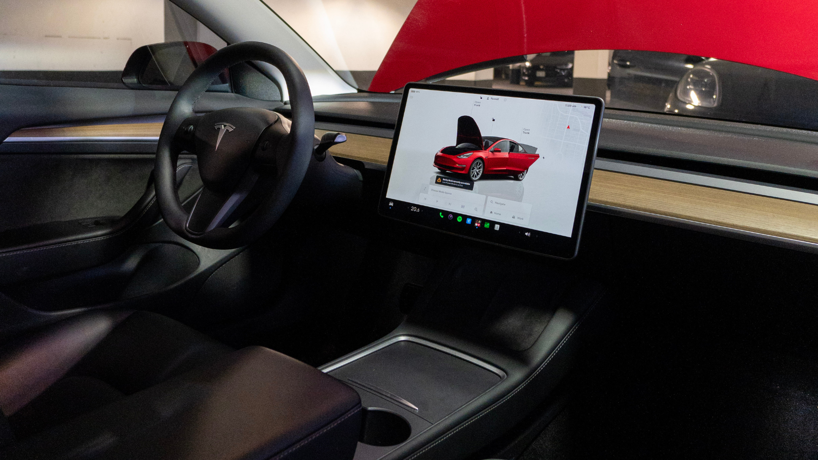 he interior of a Tesla Model 3 showing the minimalist dashboard with a large touchscreen display in the center. The screen shows a 3D graphic of a red Tesla with the front trunk open alongside a navigation map. The flat bottom steering wheel with the Tesla logo is visible on the left and the center console with a wireless charging pad and cup holder is in the foreground.