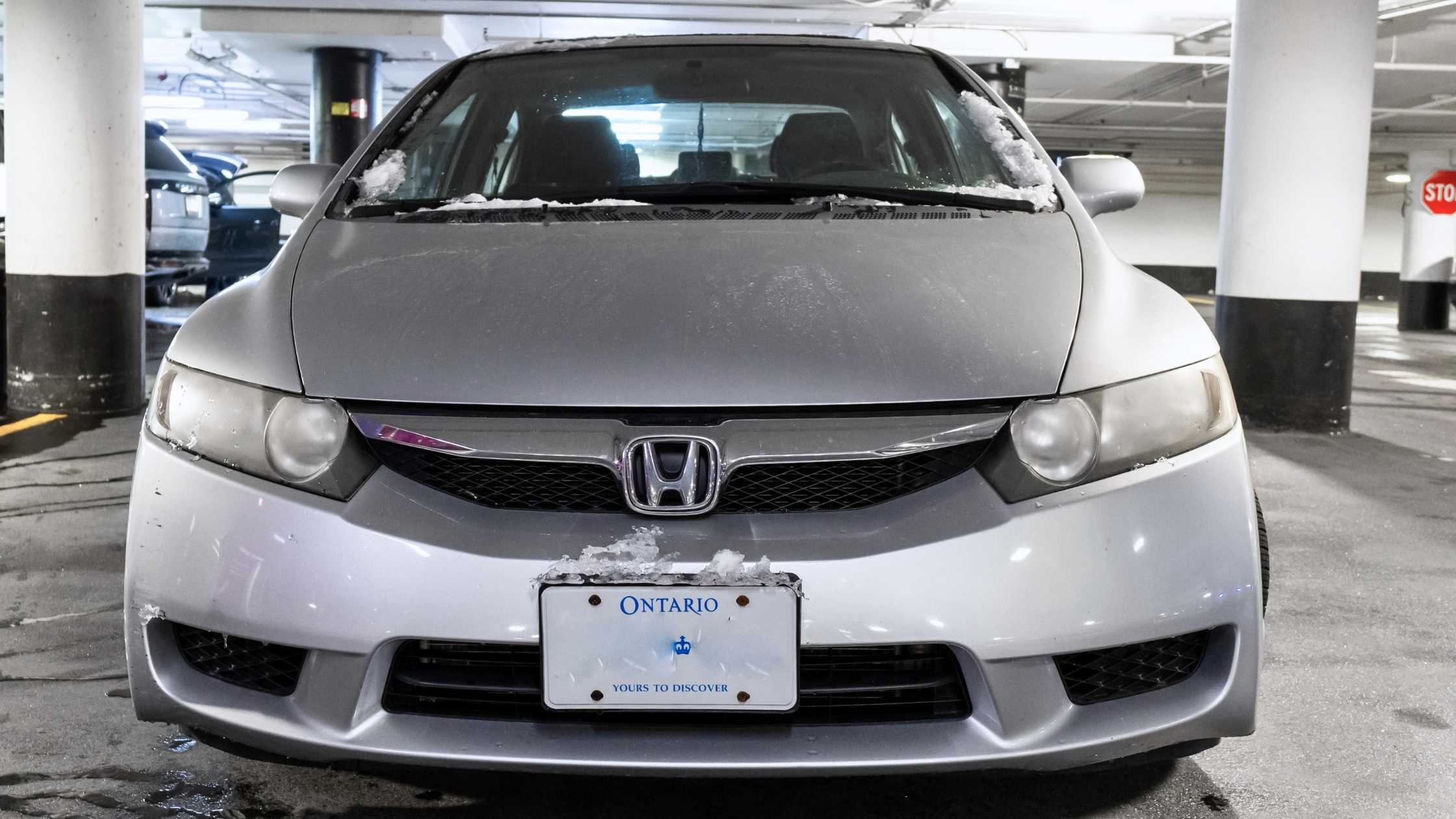 Front view of a silver Honda Civic with salt residue and snow buildup on the hood and windshield, parked inside an underground garage with visible Ontario license plate.